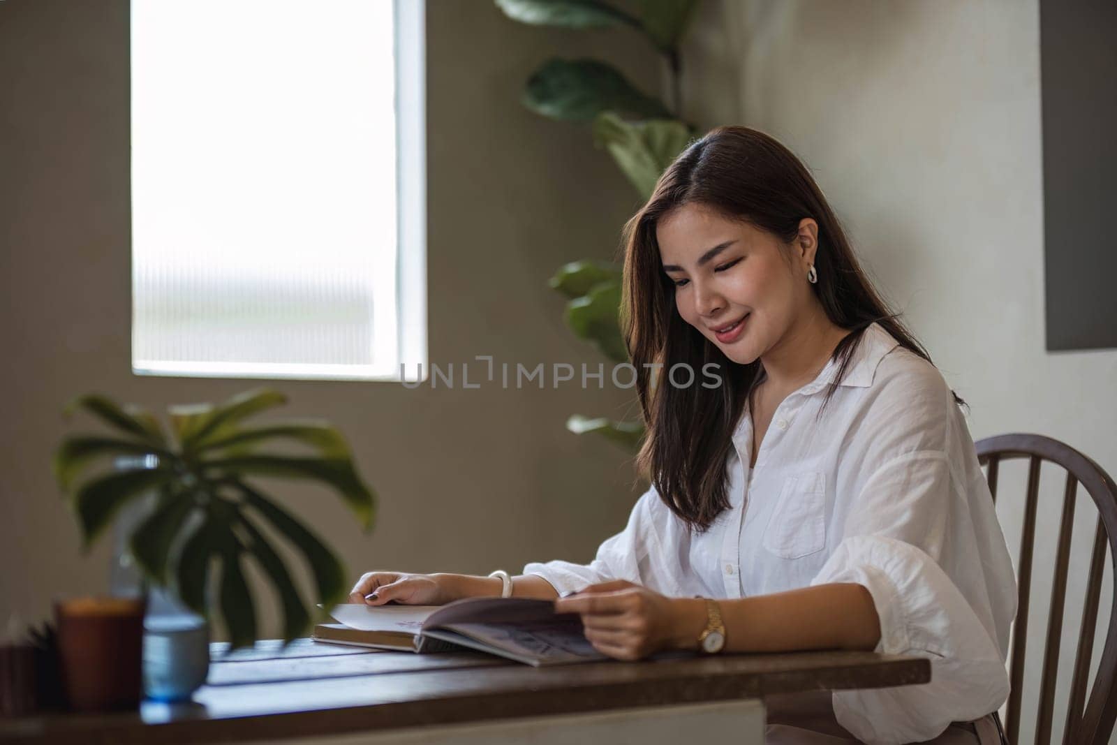 Attractive and happy young Asian girl reading a book and use laptop at a table in her minimal living room. Hobby and leisure concepts by wichayada