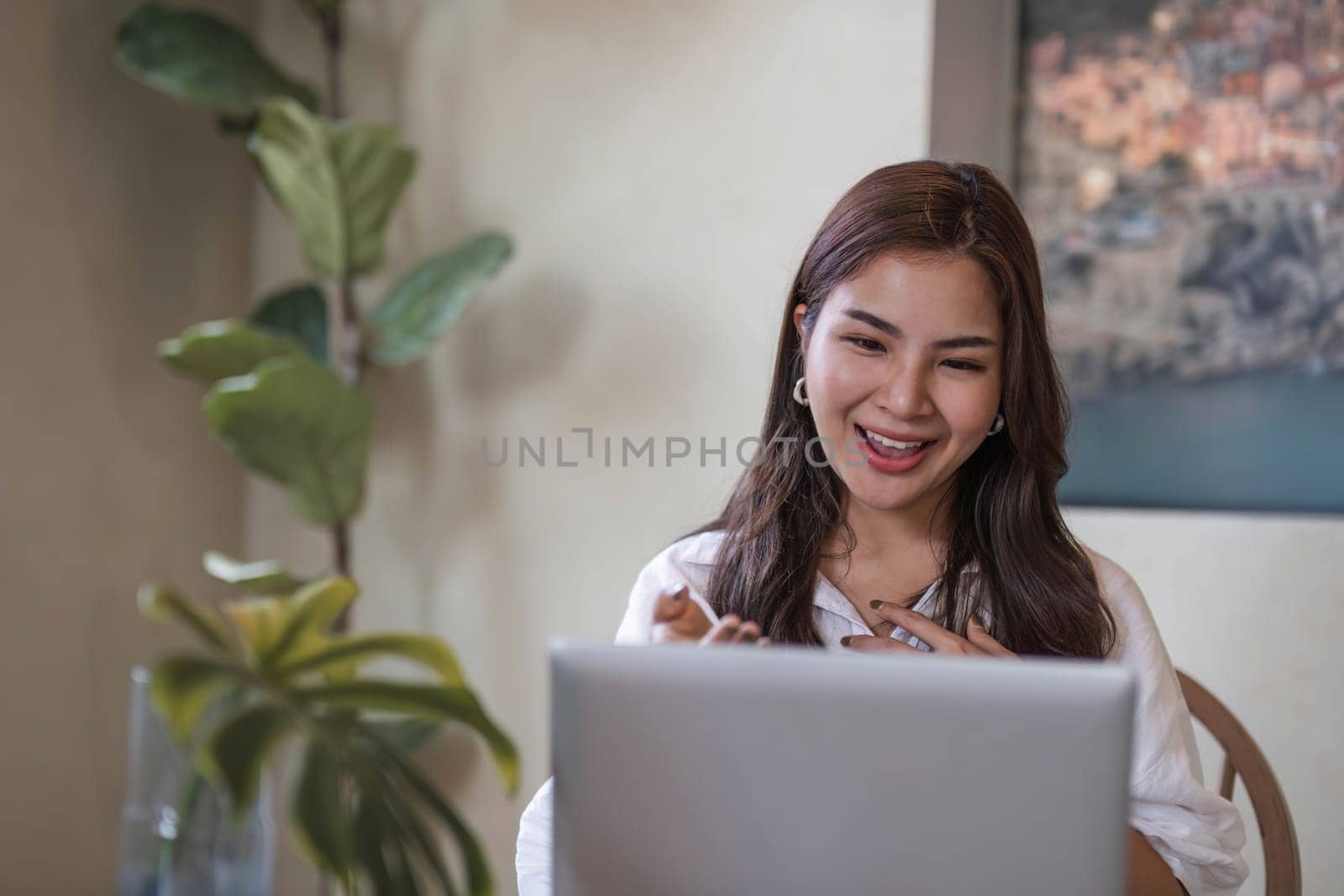 Attractive and happy young Asian girl reading a book and use laptop at a table in her minimal living room. Hobby and leisure concepts..