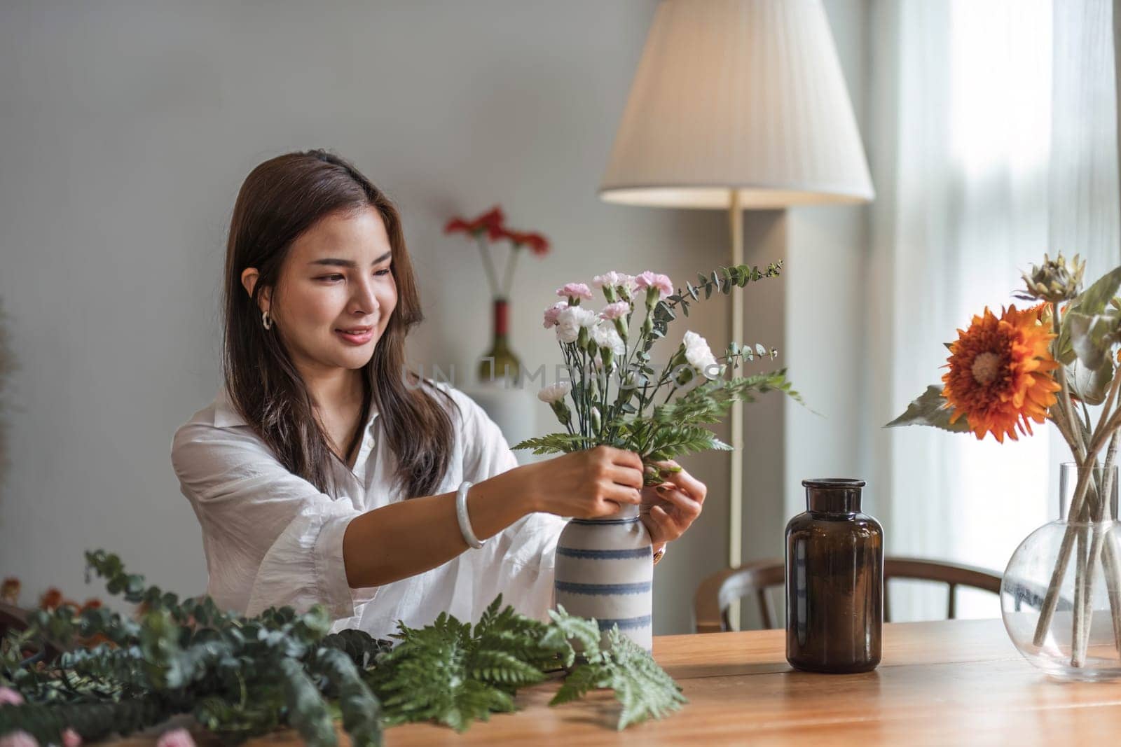 Attractive and happy young Asian woman enjoys arranging a vase with beautiful flowers in her minimal living room. Leisure and lifestyle concepts..