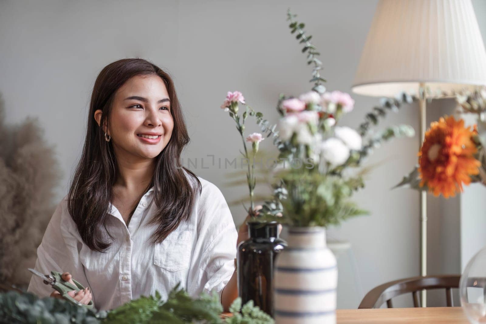 Attractive and happy young Asian woman enjoys arranging a vase with beautiful flowers in her minimal living room. Leisure and lifestyle concepts by wichayada