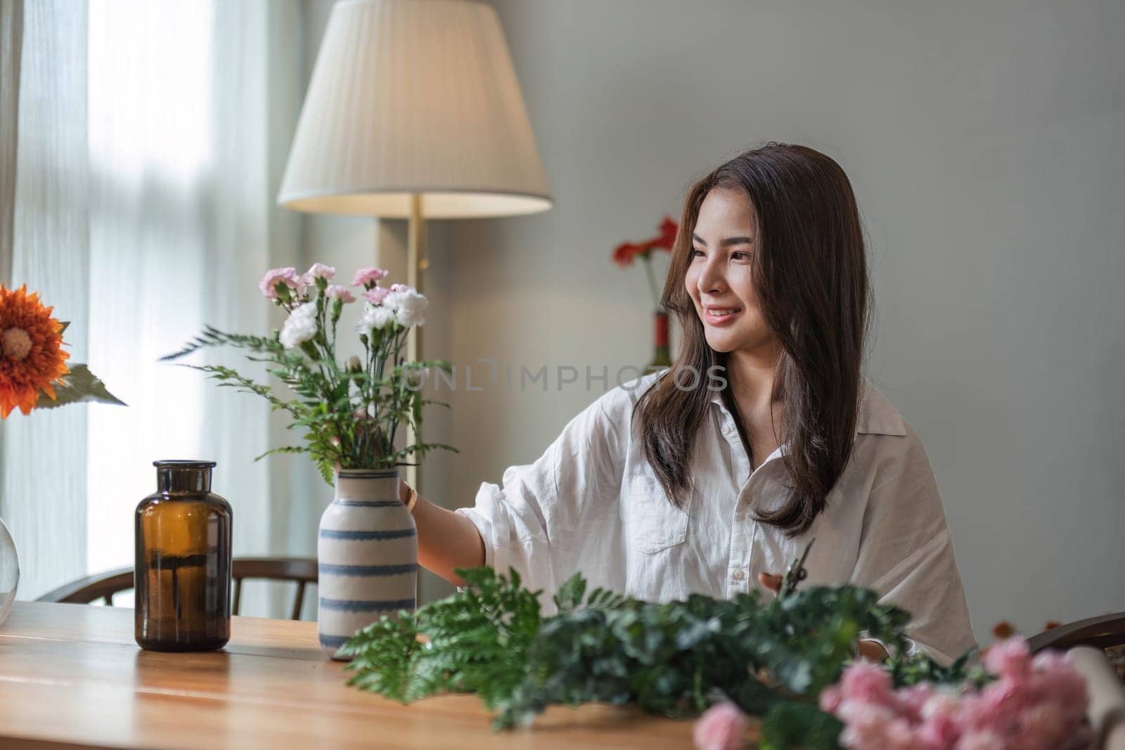 Attractive and happy young Asian woman enjoys arranging a vase with beautiful flowers in her minimal living room. Leisure and lifestyle concepts..