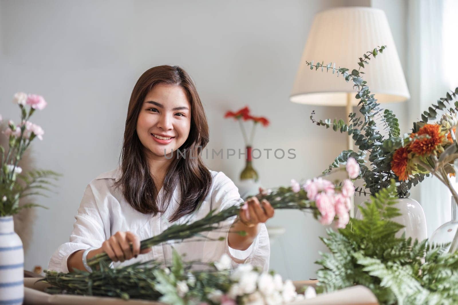 Attractive and happy young Asian woman enjoys arranging a vase with beautiful flowers in her minimal living room. Leisure and lifestyle concepts by wichayada