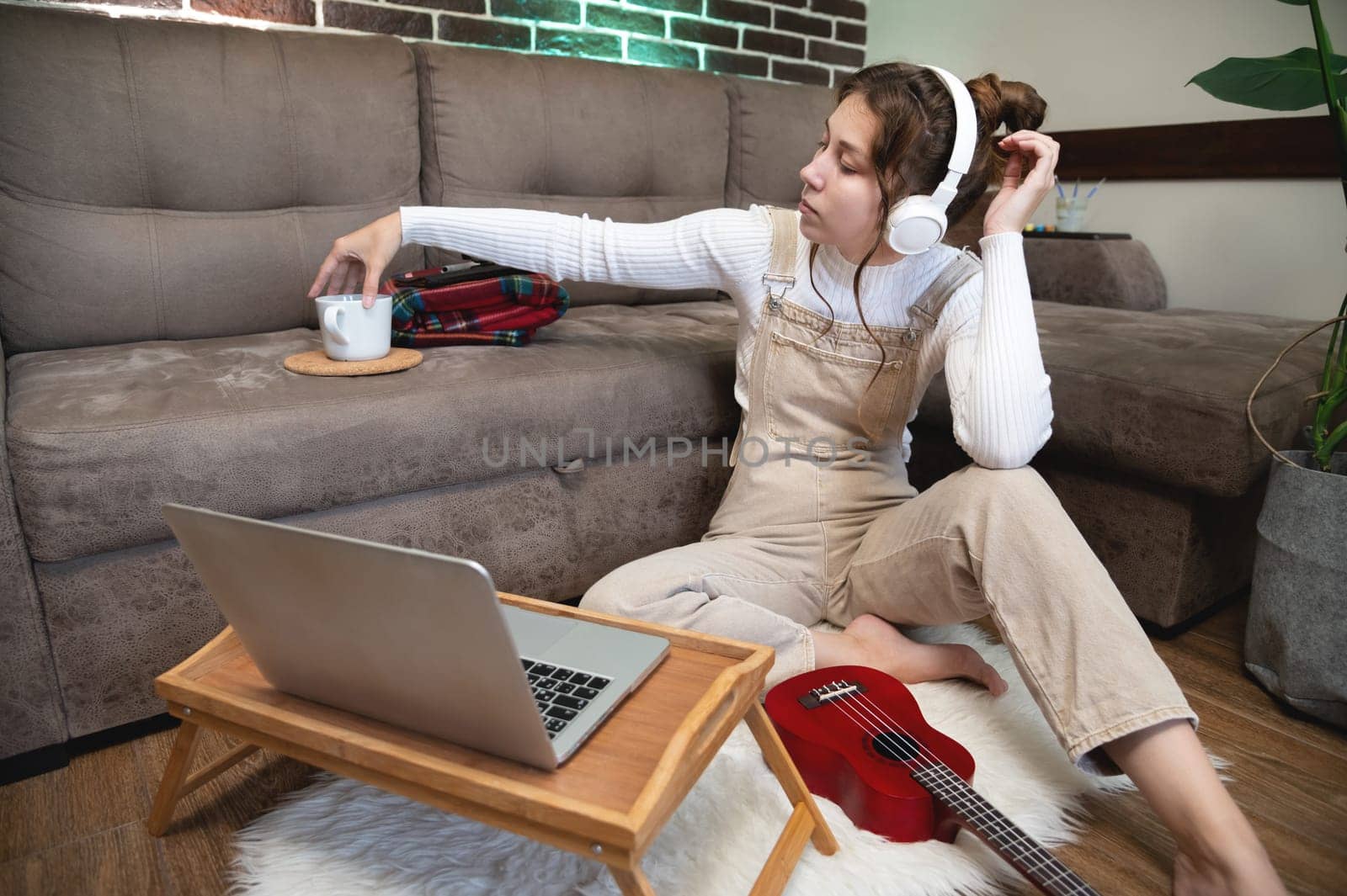 Image of a smiling caucasian young woman drinking hot tea and sitting next to a laptop on the floor, relaxing at home. Thoughtful professional working from home or student looking away while on a call by yanik88