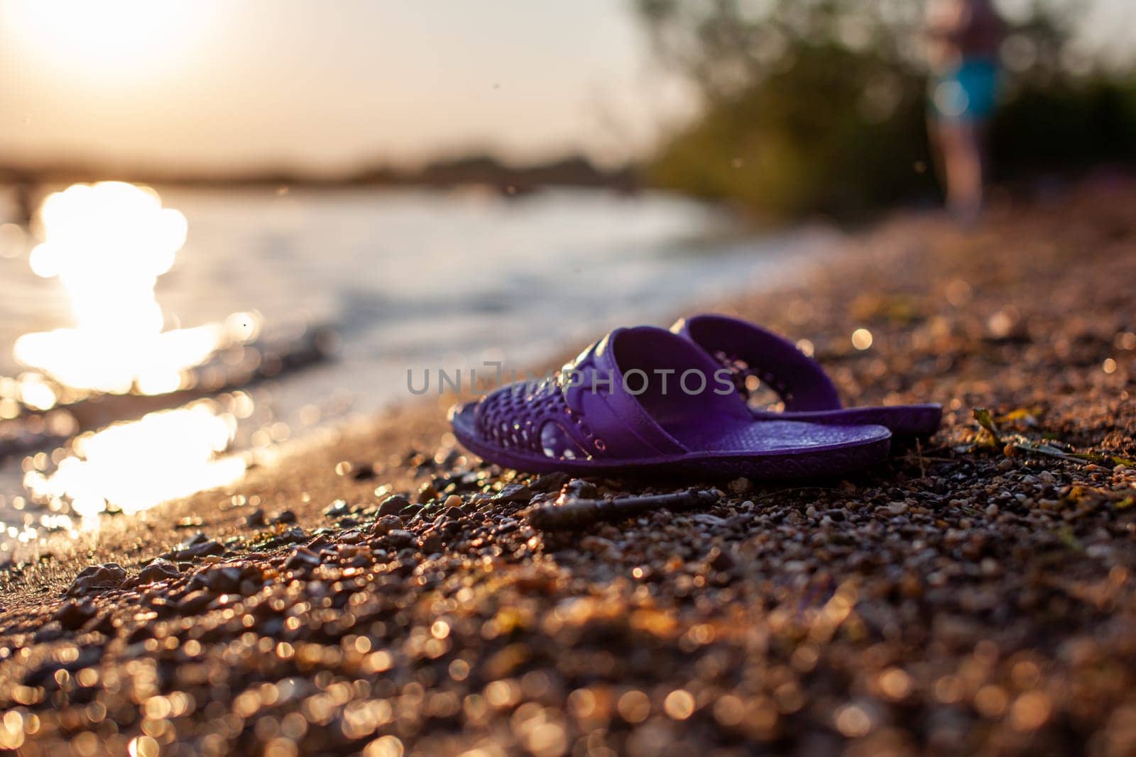 Close-up of rubber slippers on the shore of a lake or river made of small stones against a colorful sunset background. A beautiful place in nature for family holidays and swimming.