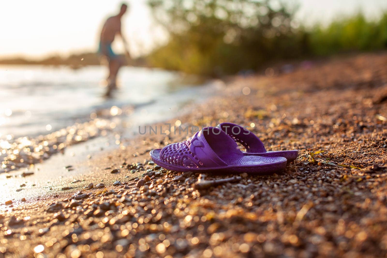 Close-up of rubber slippers on the shore of a lake or river made of small stones against a colorful sunset background. A beautiful place in nature for family holidays and swimming.