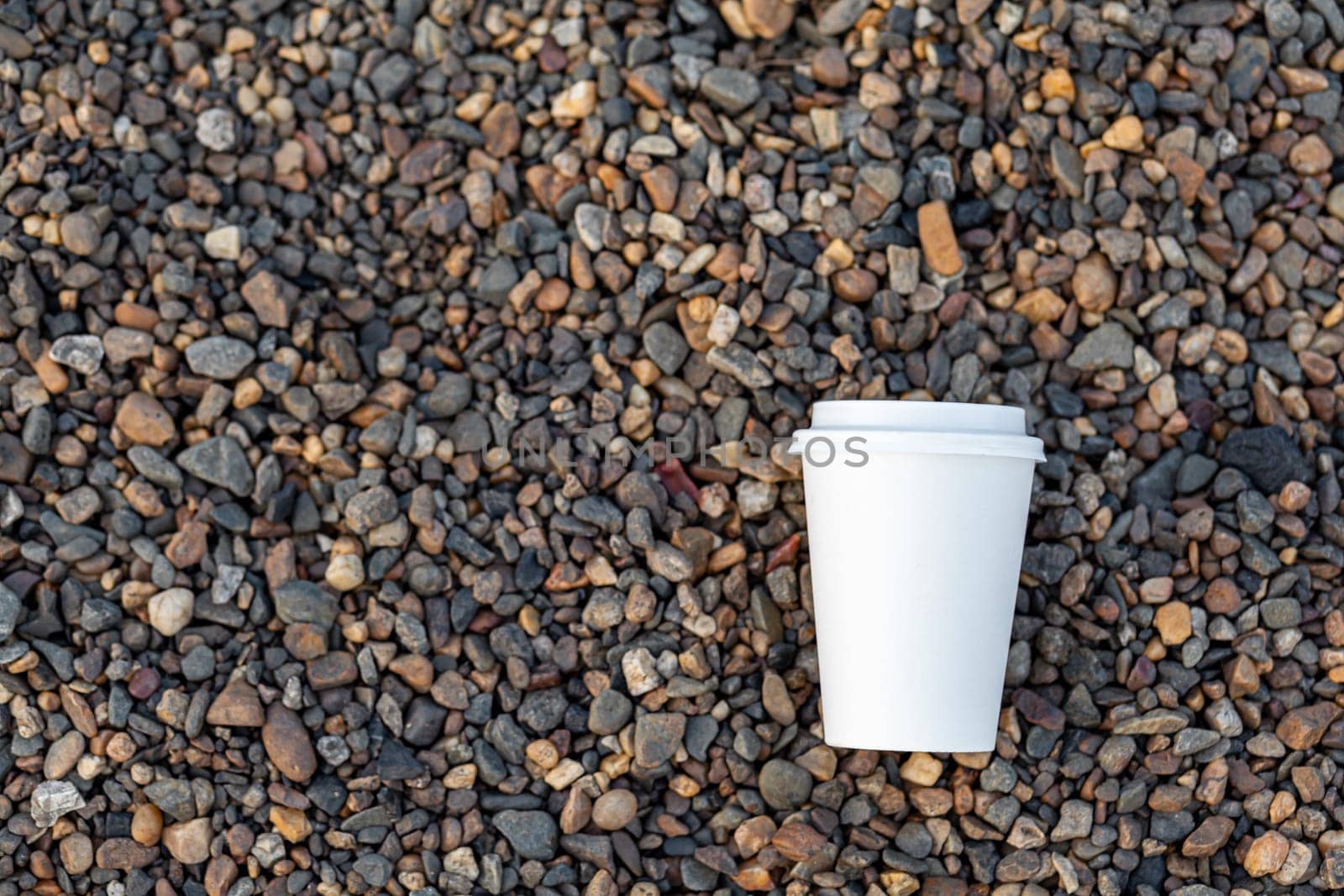 A white paper cup of coffee or tea lies on the rocky shore. A mug of hot drink on a pebble. Beautiful rocky background. free copy space.