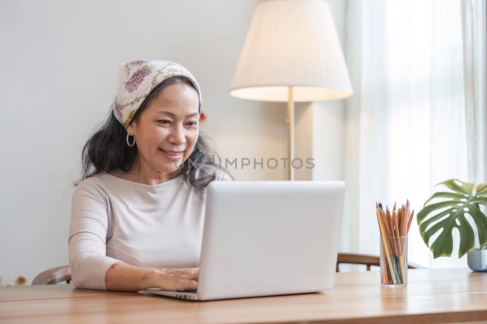 Attractive and relaxed 60s mature Asian woman in casual clothes use laptop at a table in her living room. Leisure and lifestyle concepts.