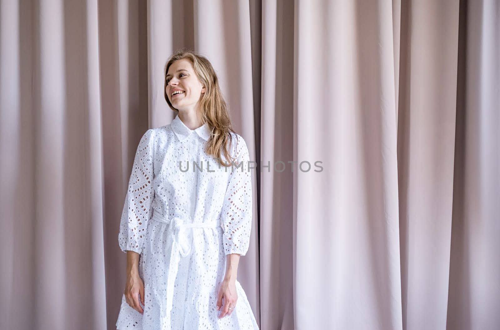Beautiful caucasian woman in white summer fashion dress standing on beige curtain background, looking away. Pastel colors