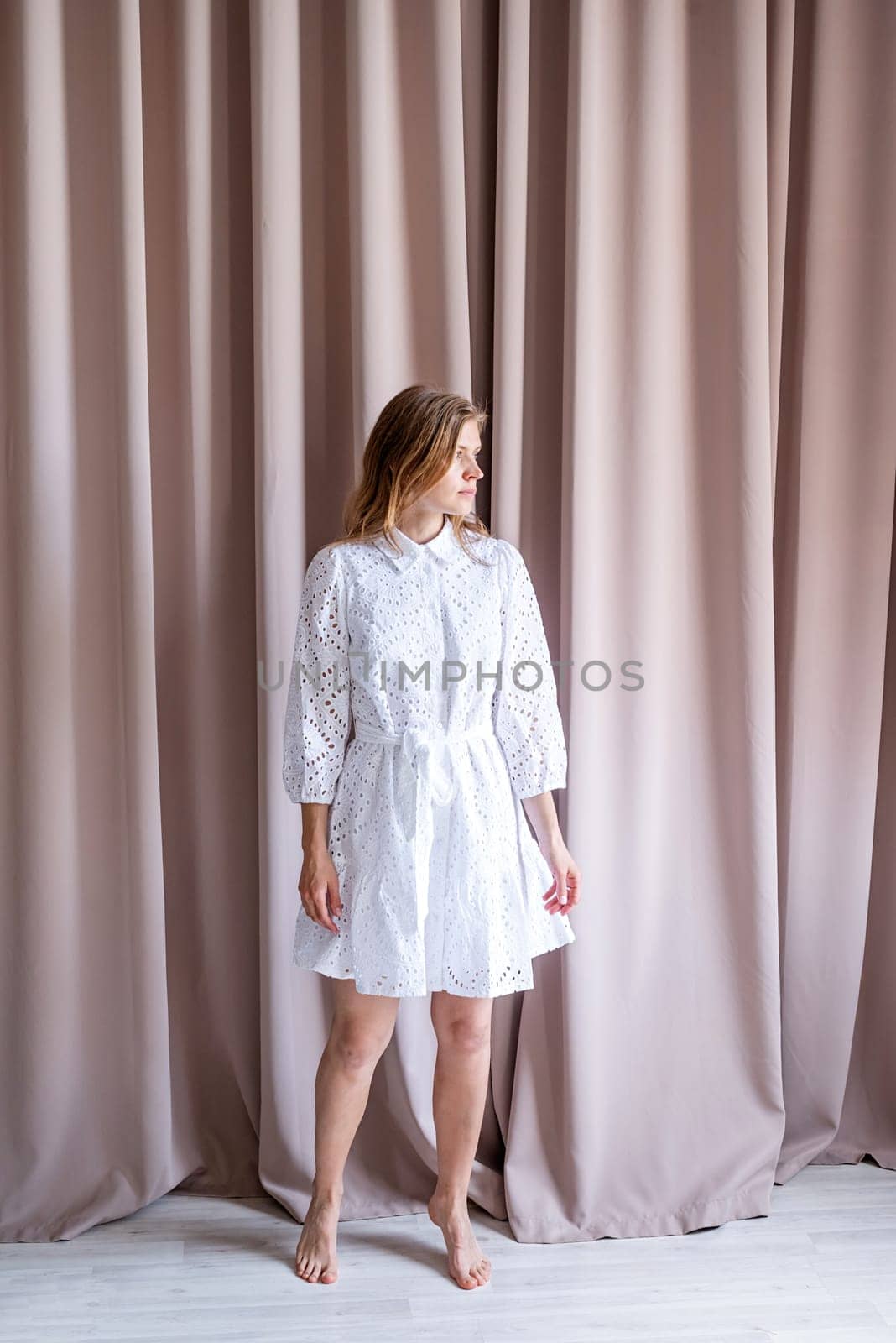 Beautiful caucasian woman in white summer fashion dress standing barefoot on beige curtain background, looking away. Pastel colors