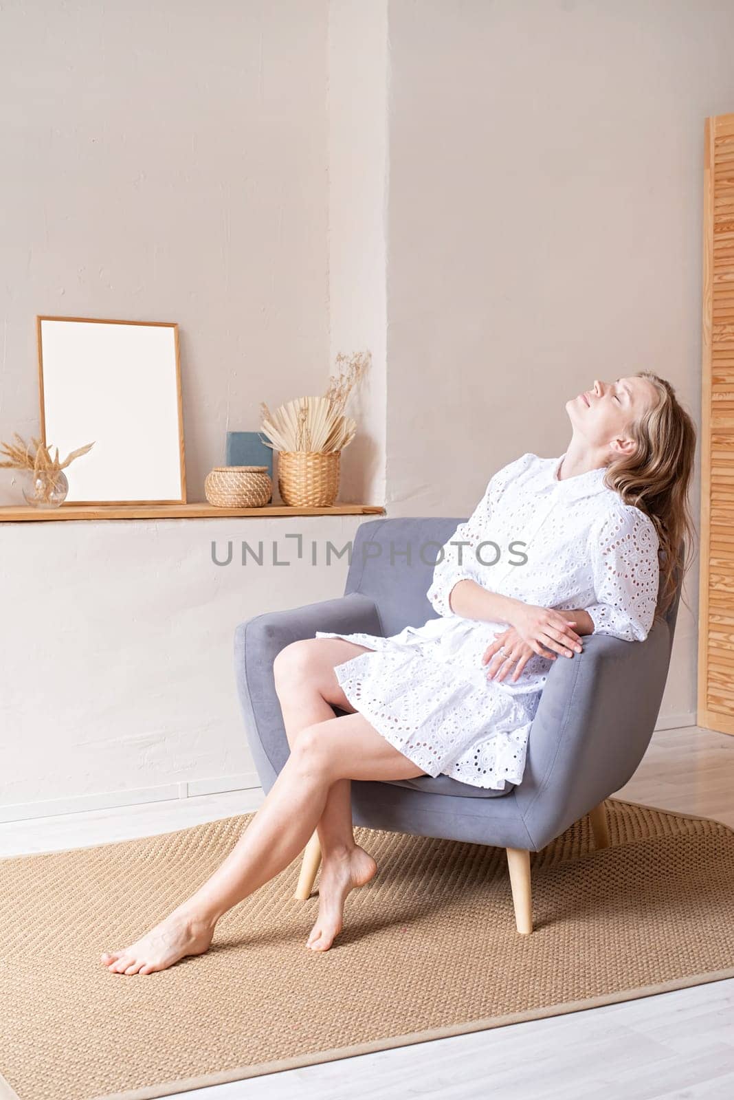 Beautiful woman in fashion dress sitting on soft chair barefoot on white wall background by Desperada