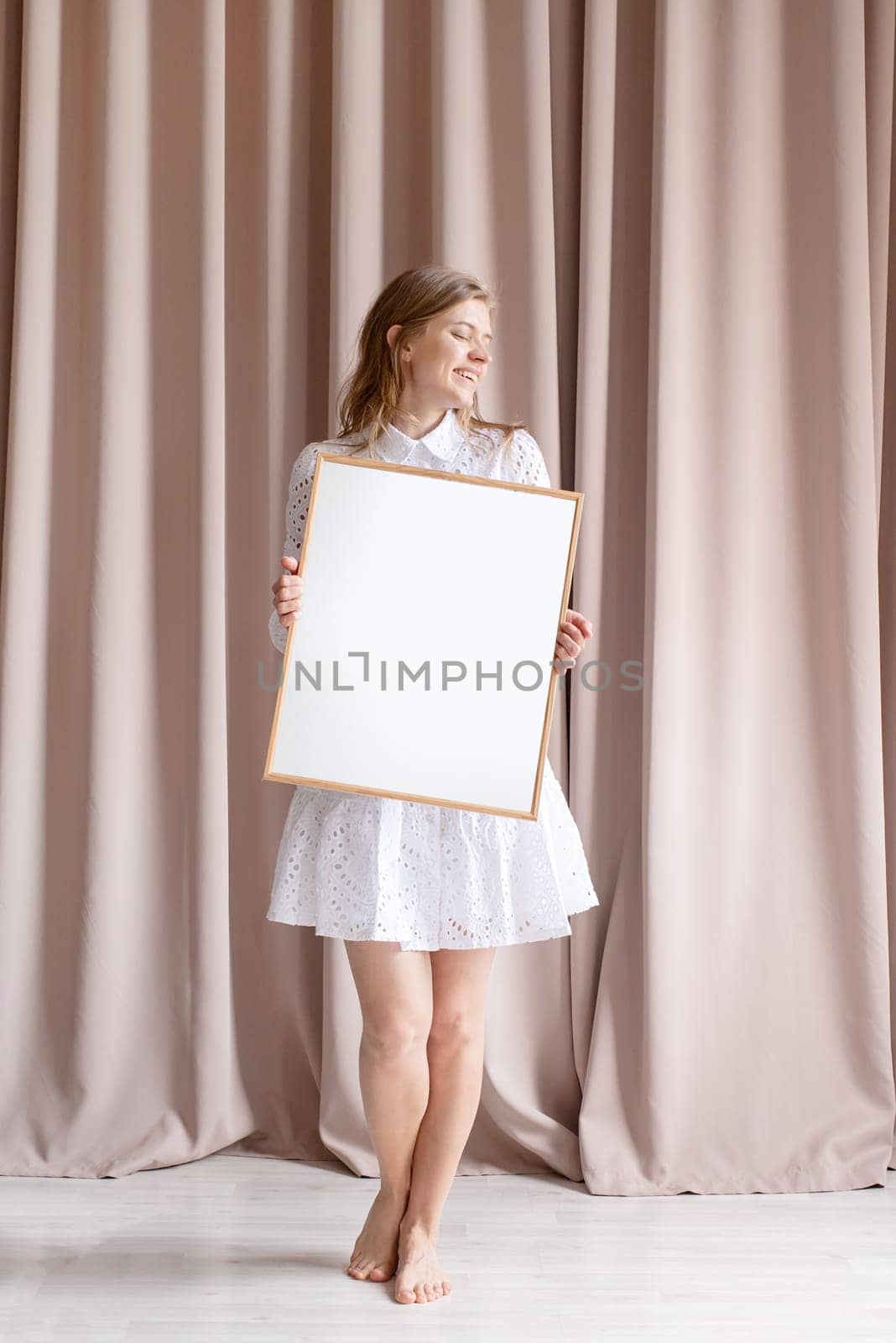 barefoot woman in beautiful white dress holding blank frame, beige curtain background, mockup design