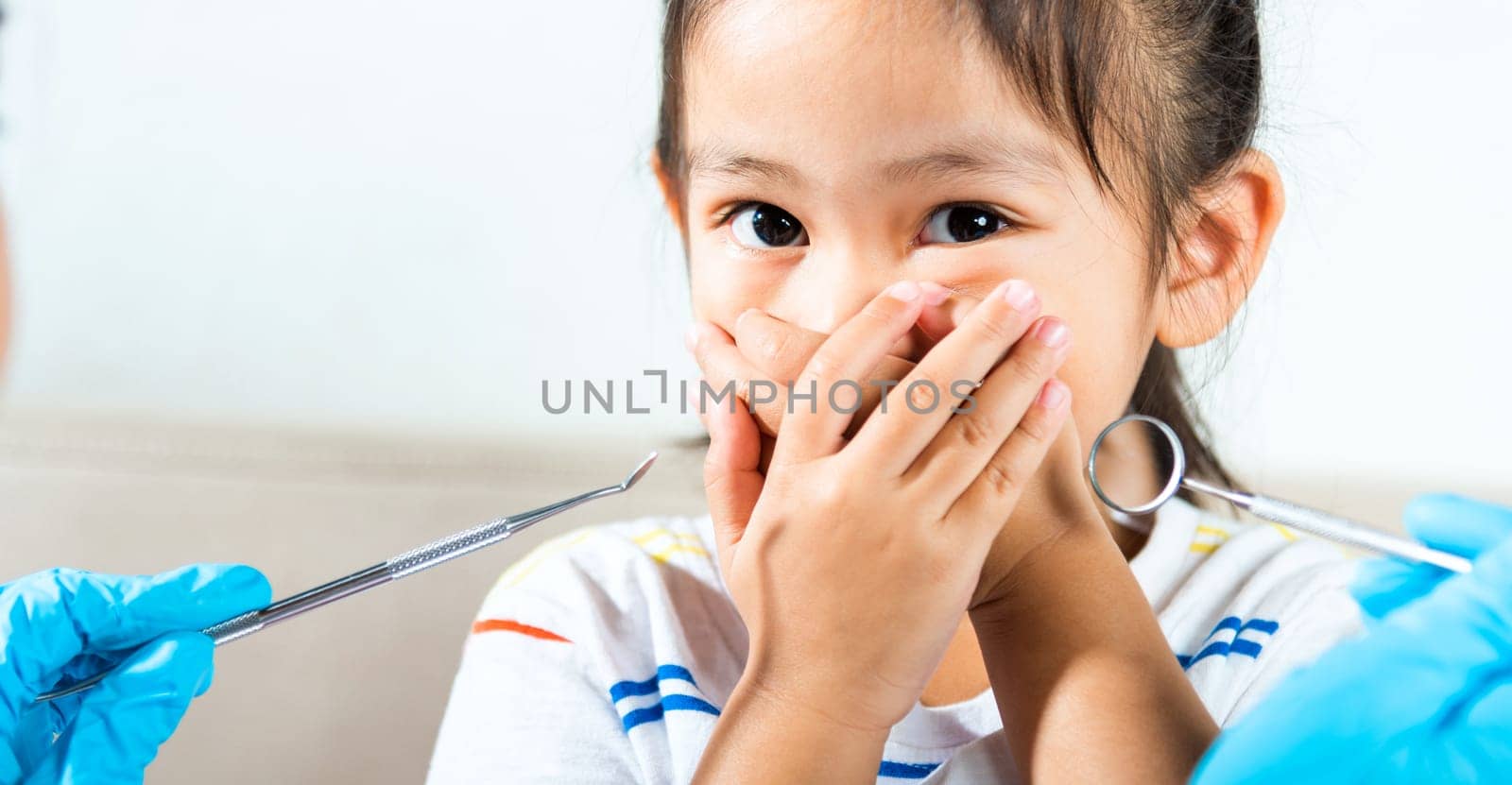 Dental kid examination. Doctor examines oral cavity of child uses mouth mirror to check teeth cavity but little girl cover mouth no need to checking she afraid, scared and closes his mouth with hands