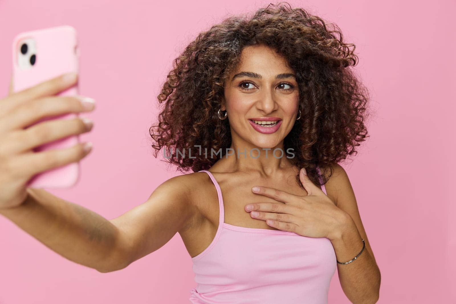Woman blogger holding phone video call takes selfies, with curly hair in pink smile t-shirt and jeans poses on pink background, copy space, technology and social media, online. High quality photo