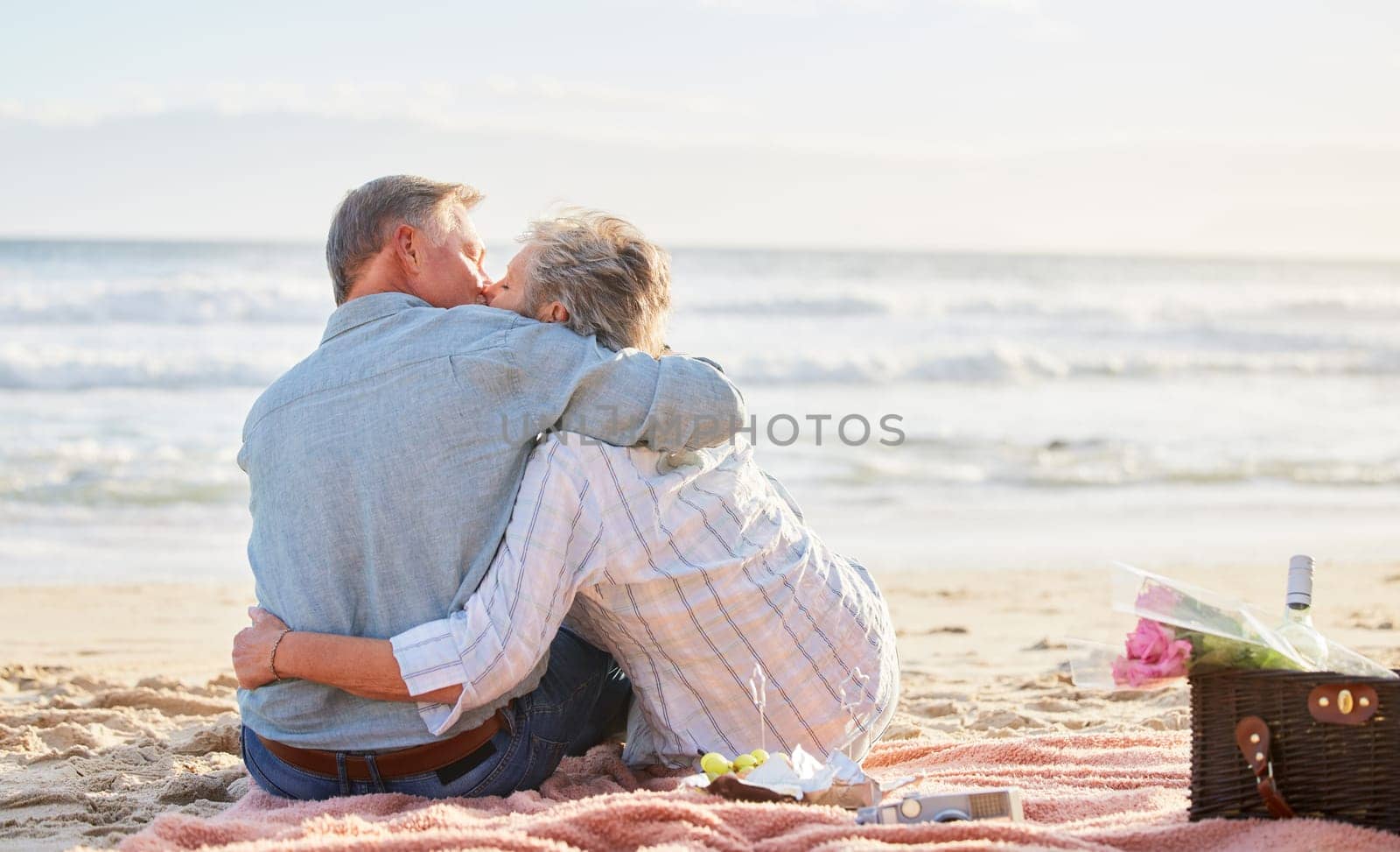 Senior couple, beach picnic and kiss with hug, romance and happiness in summer for anniversary celebration. Elderly man, old woman and basket for food, wine and outdoor for sunshine, waves and love.