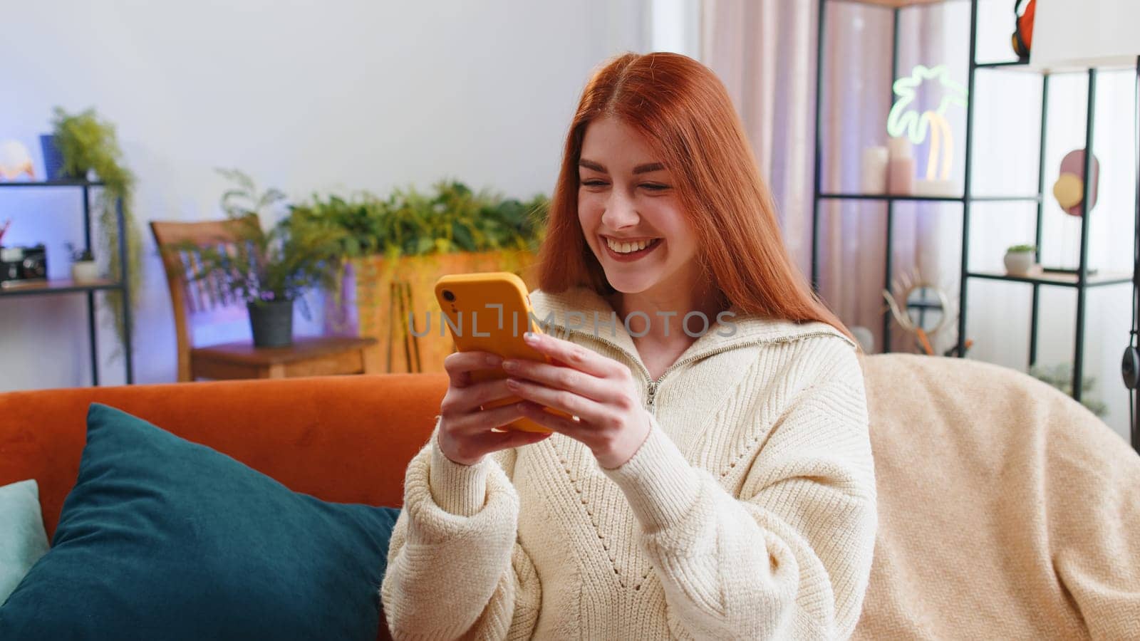 Happy young woman use mobile smartphone typing browsing say Wow yes found out great big win good news celebrate lottery jackpot doing winner gesture. Redhead girl at home in room sitting on sofa