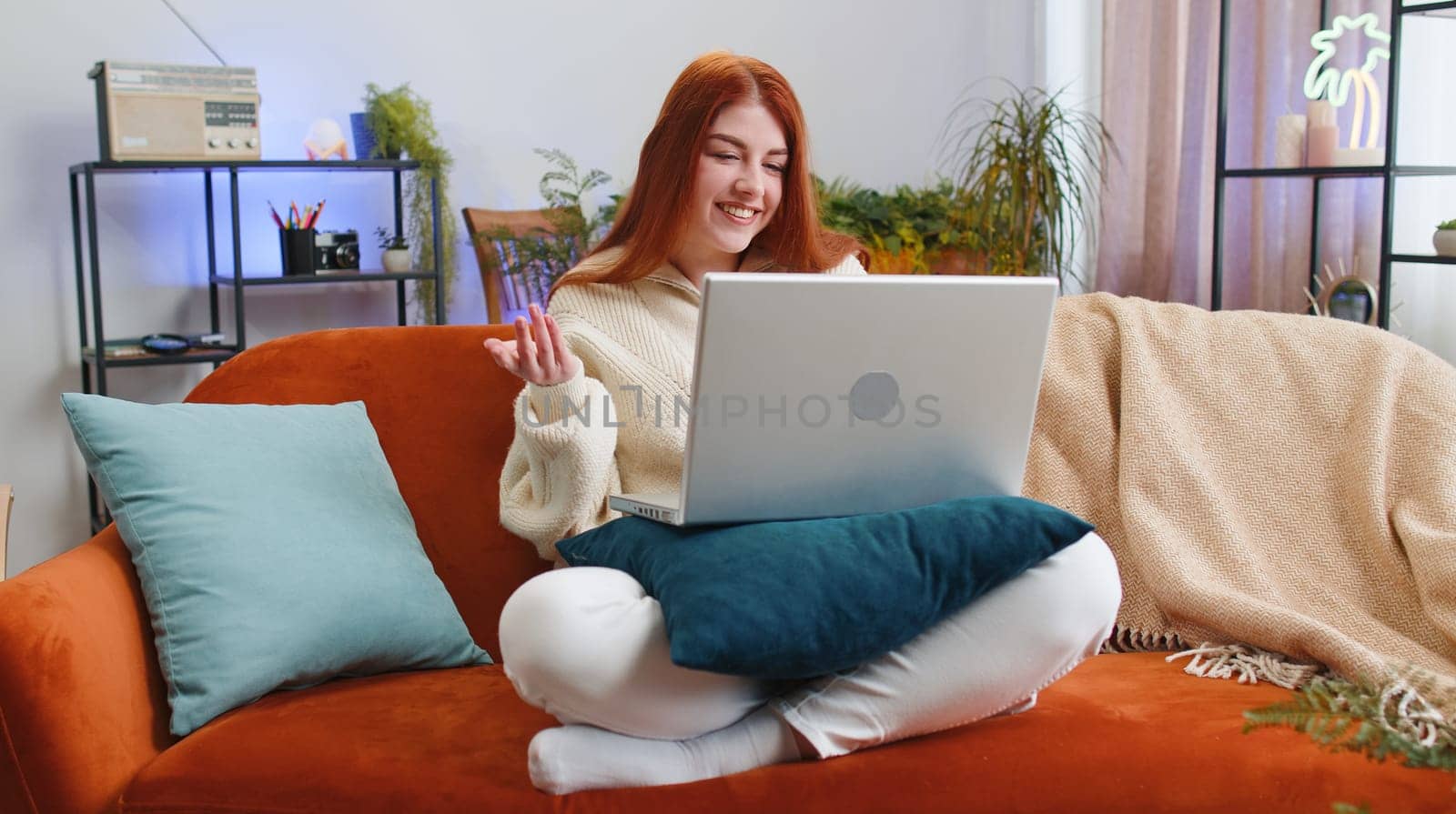 Young woman sitting on couch looking at camera, making video webcam conference call with friends or family, enjoying pleasant conversation. Portrait of redhead girl laughing waving hello at home room