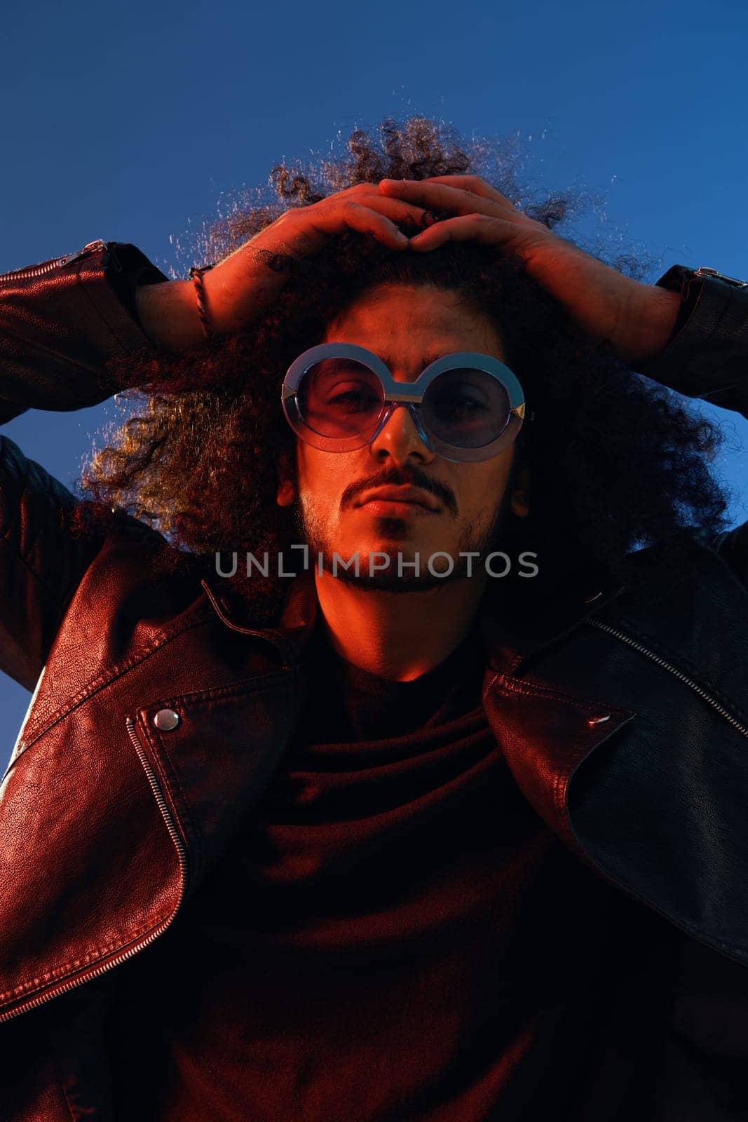 Portrait of a stylish man with curly hair with glasses smile on blue background multinational, color light, black leather jacket trend, modern concept. High quality photo