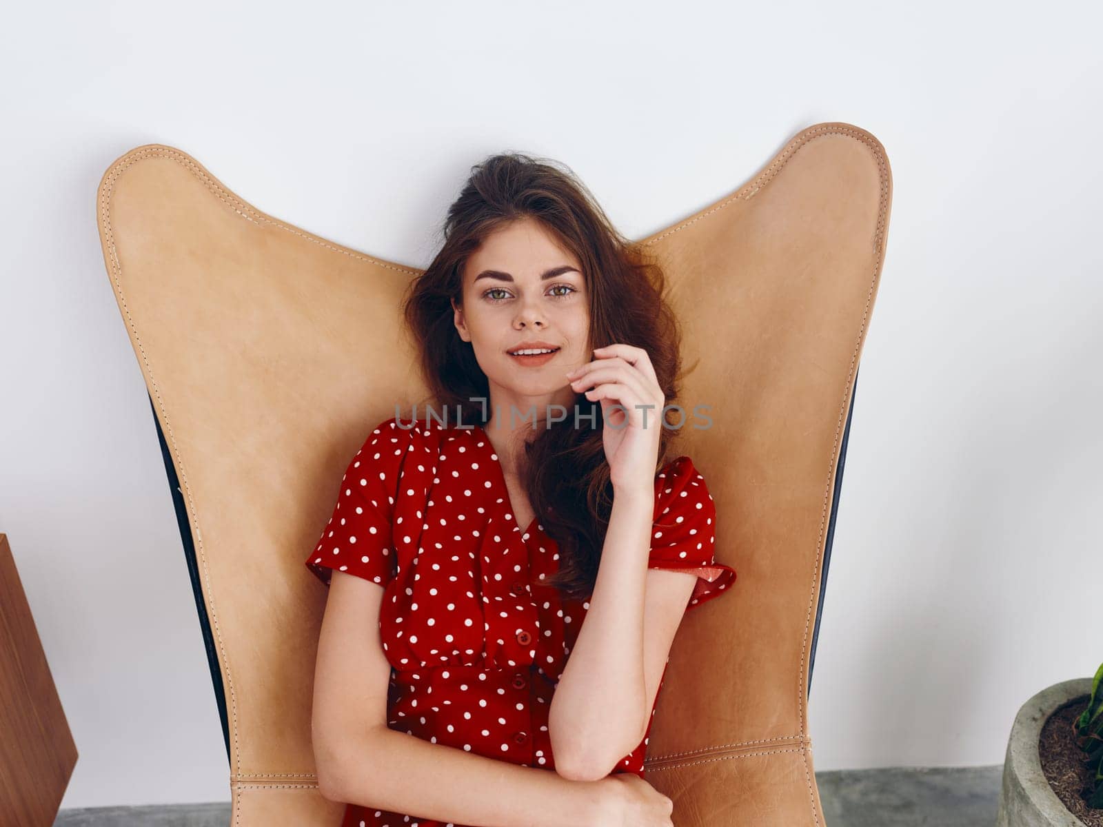 Woman sitting in leather armchair smile with teeth Lifestyle red polka dot dress, relaxing at home stylish modern interior with white walls. High quality photo