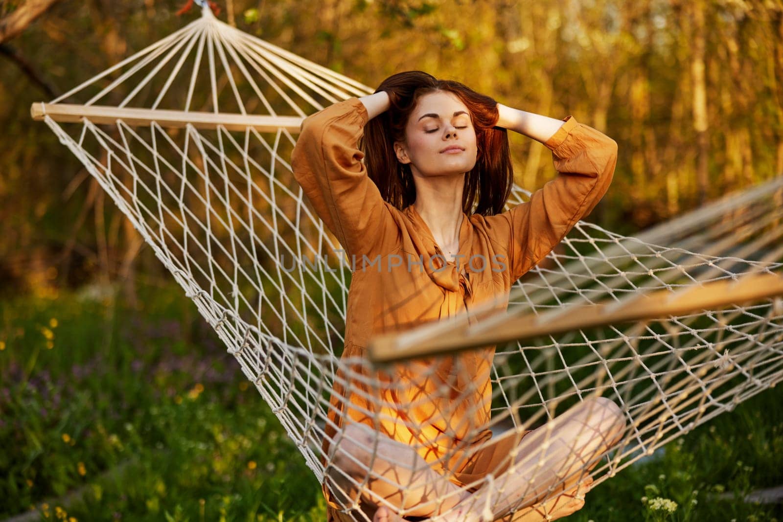 woman is sitting in a mesh hammock in nature, relaxing and enjoying the rays of the setting sun, straightening her hair with her hands, closing her eyes on a warm summer day. Horizontal photo by Vichizh
