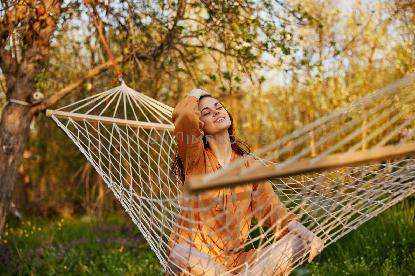 a happy woman in a long orange dress is resting lying in a hammock at the dacha, straightening her long hair and looking away, illuminated by the summer sun during sunset by Vichizh