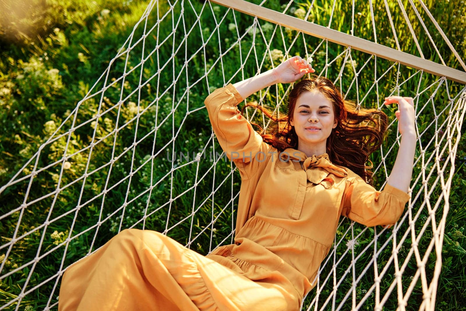 a beautiful, elegant woman lies in a long orange dress on a mesh hammock resting in nature, illuminated by the warm sunset light, happily smiling, straightens her hair with her hands by Vichizh