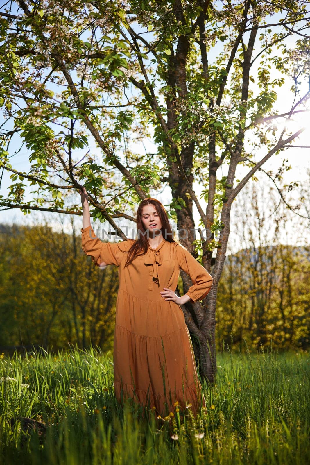 a sweet, attractive woman with long red hair is standing in the countryside near a flowering tree in a long orange dress and touching a branch with her hand looks at the camera by Vichizh
