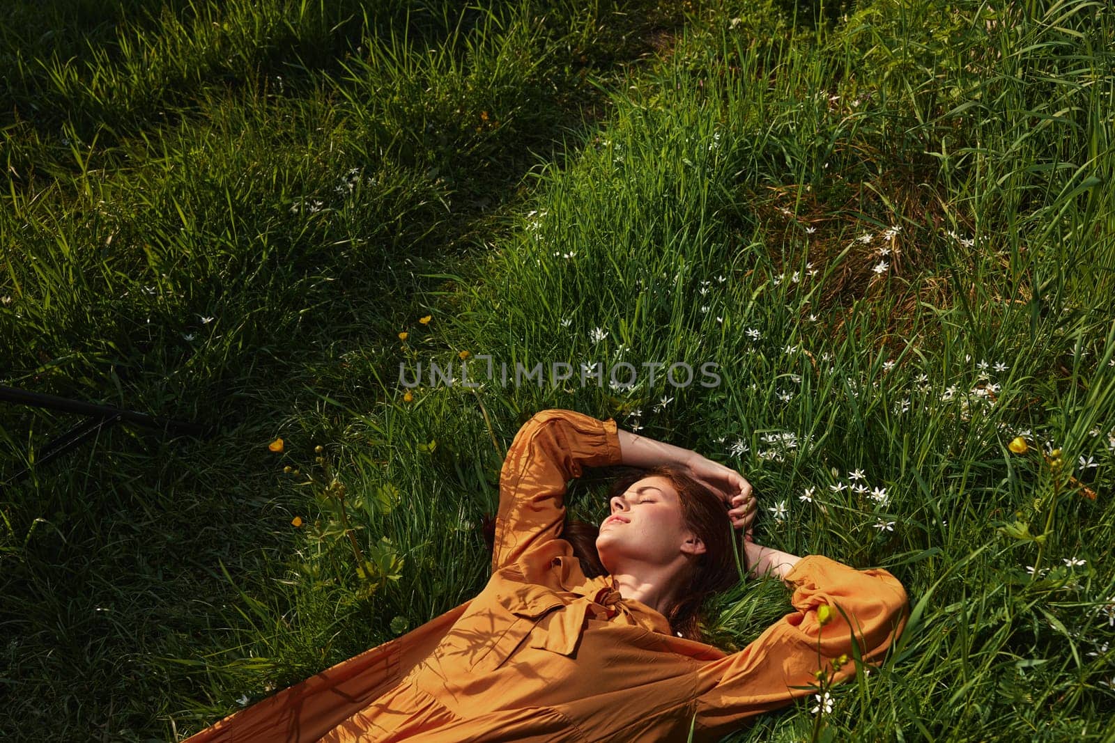 horizontal photo of a happy, relaxed woman, resting lying in the grass, in a long orange dress, with closed eyes and a pleasant smile on her face, enjoying harmony with nature and recuperating by Vichizh