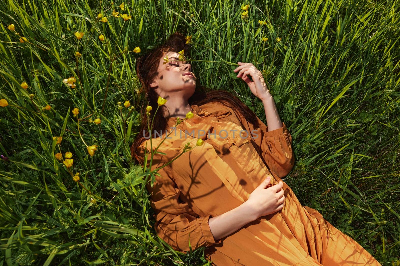 a calm woman with long red hair lies in a green field with yellow flowers, in an orange dress with her eyes closed, with a pleasant smile on her face, enjoying peace and recuperating. High quality photo
