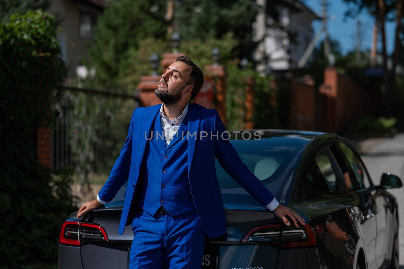 Caucasian bearded man in a blue suit stands near a black car in the countryside in summer