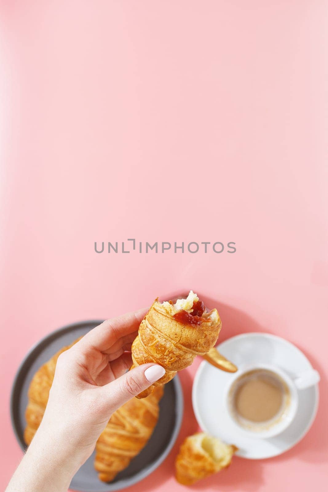 A woman's hand holds a croissant over a plate with fresh puff croissants, coffee and berry jam on a pink background. copy space.