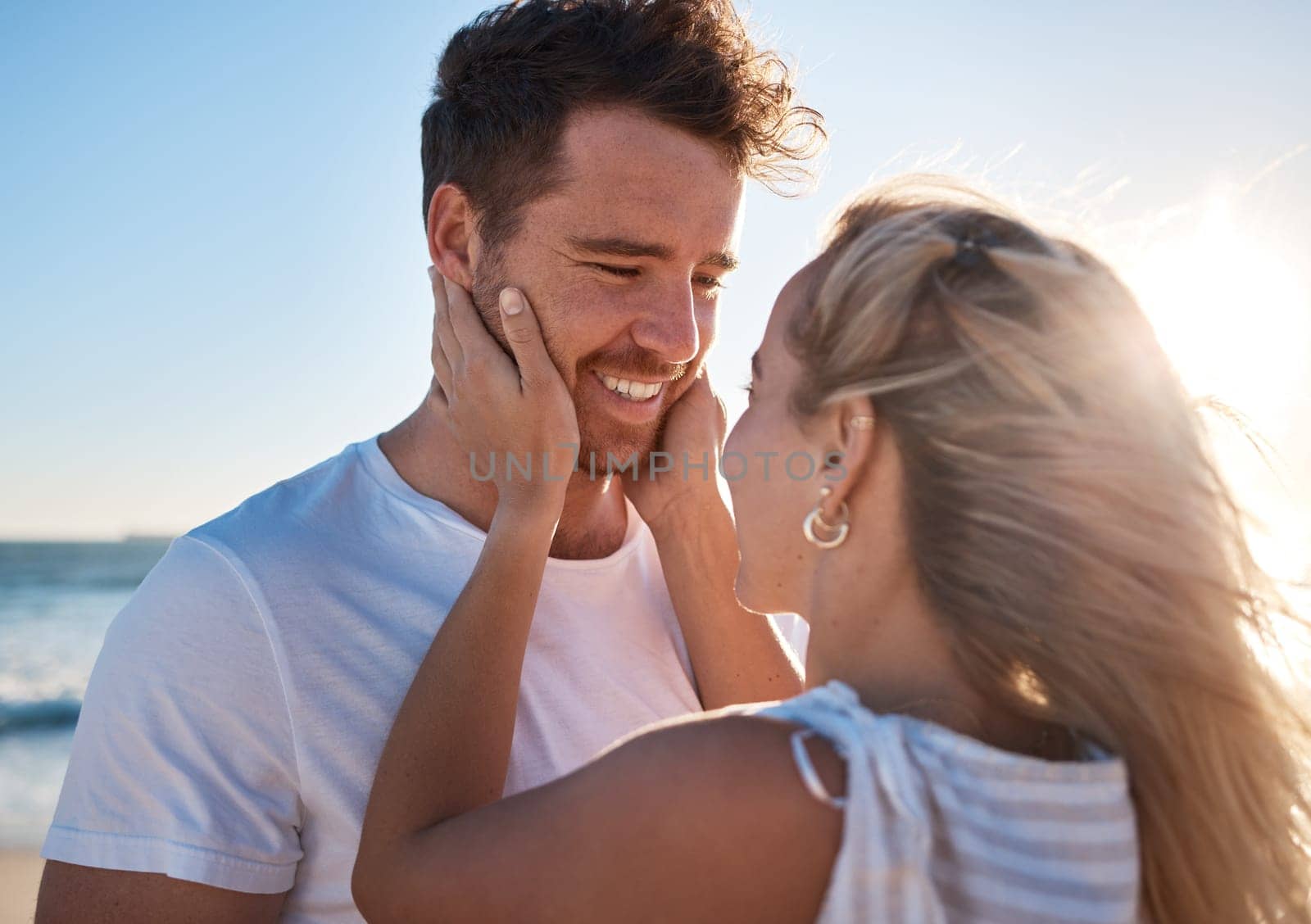 Woman, man and touch face, love and happy together at the beach, travel and romance outdoor for bonding. Couple on adventure by the ocean, embrace with lens flare and care while spending quality time.