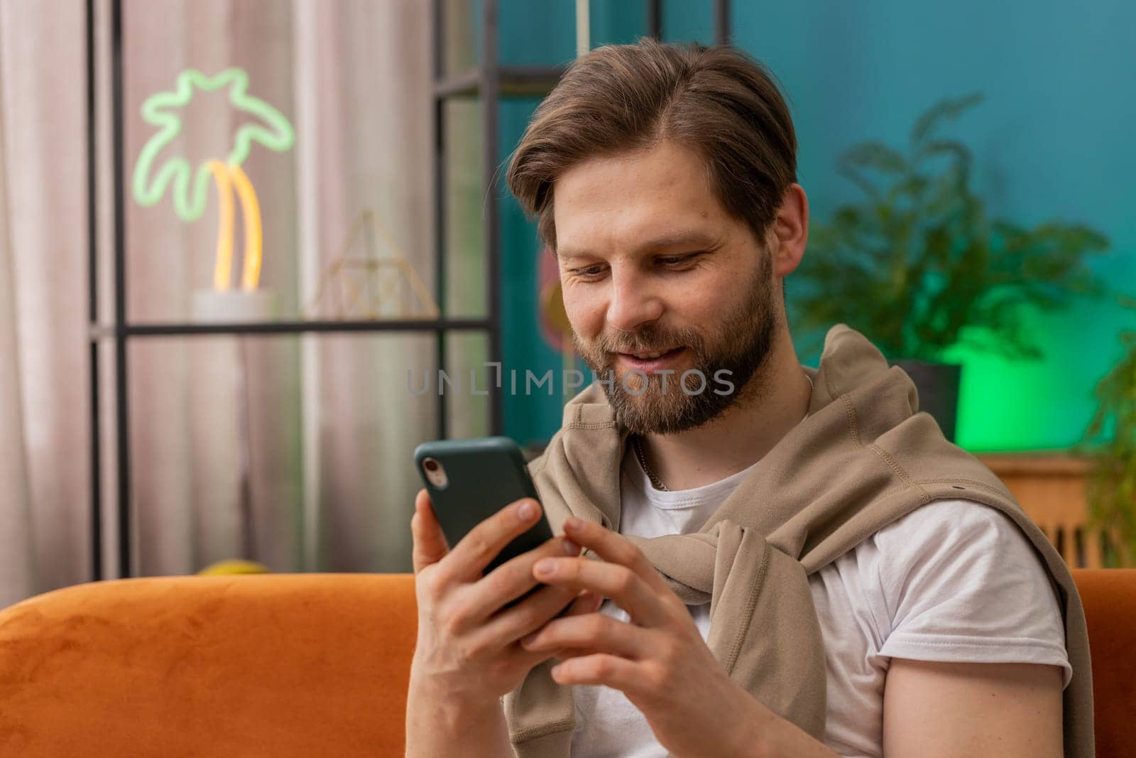Caucasian man sitting on couch uses mobile phone smiles at home living room apartment. Young guy texting share messages content on smartphone social media applications online, watching relax movie