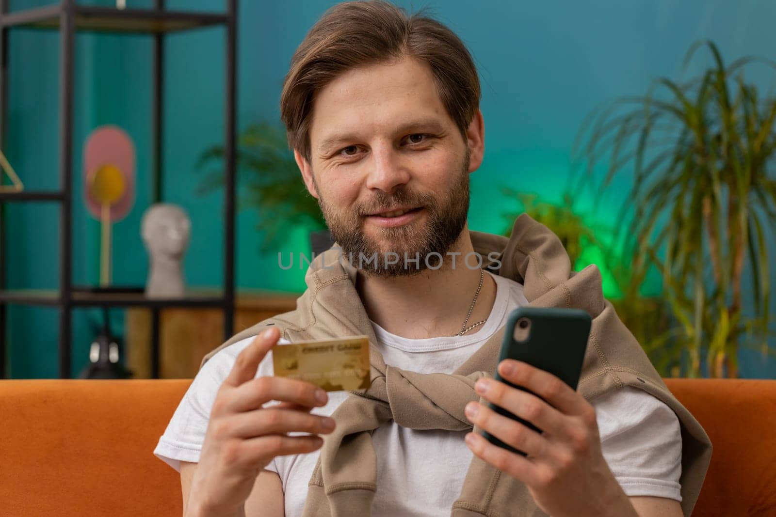 Smiling man using credit bank card and smartphone while transferring money, purchases online shopping cashless order food delivery at home apartment indoors. Happy young guy sitting in room on couch