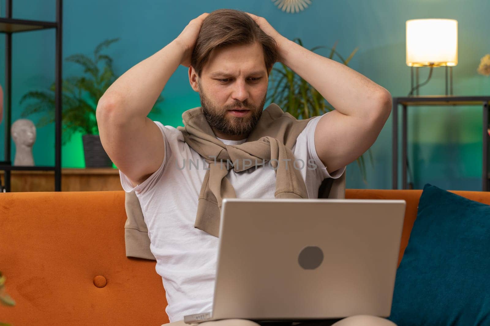 Displeased Caucasian man use laptop notebook typing browsing working, loses becoming surprised sudden lottery results bad news fortune loss fail computer virus. Young guy at home in room on couch