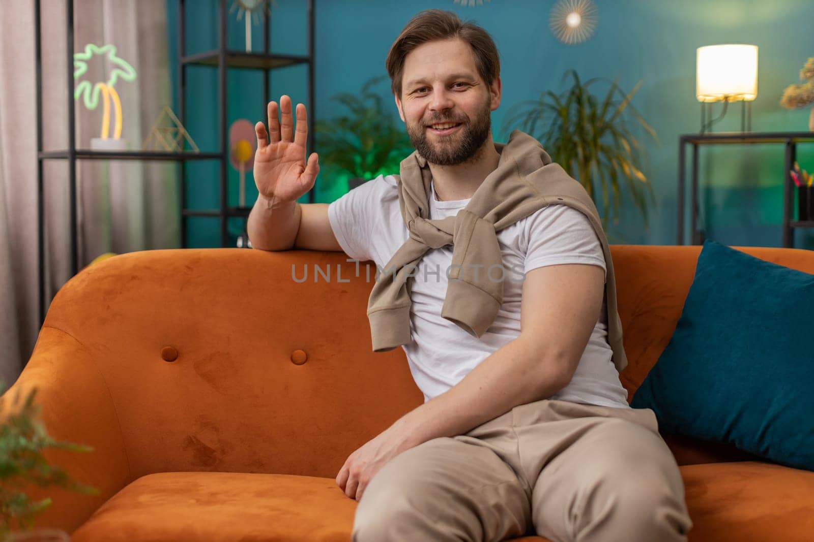 Hello. Caucasian handsome man smiling friendly at camera and waving hands gesturing hello, hi, greeting or goodbye, welcoming with hospitable expression at home. Guy sitting on sofa in living room
