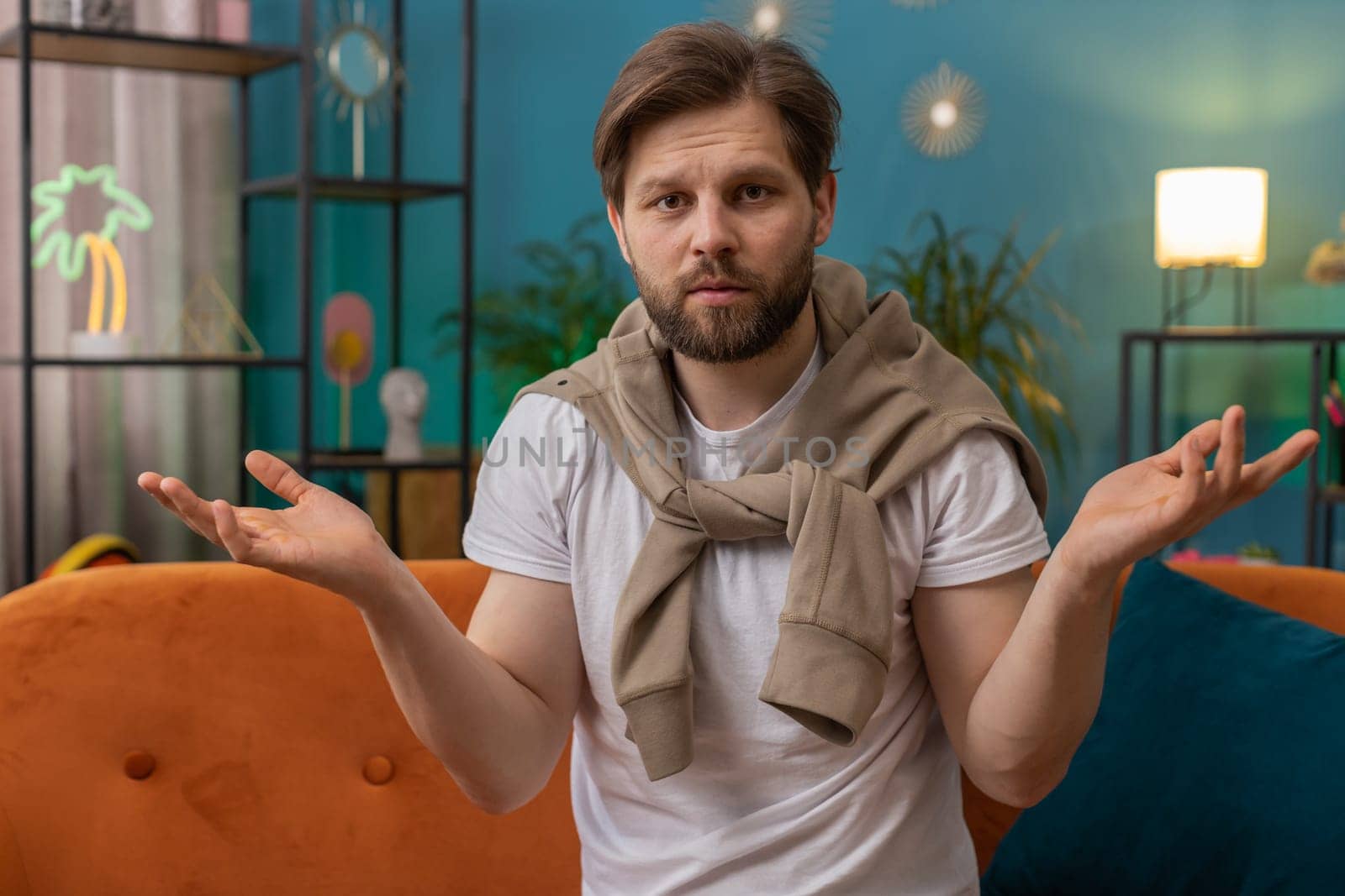 What. Why. Caucasian man raising hands in indignant expression asking reason of failure demonstrating disbelief irritation by troubles at home apartment. Young guy in living room sitting on sofa