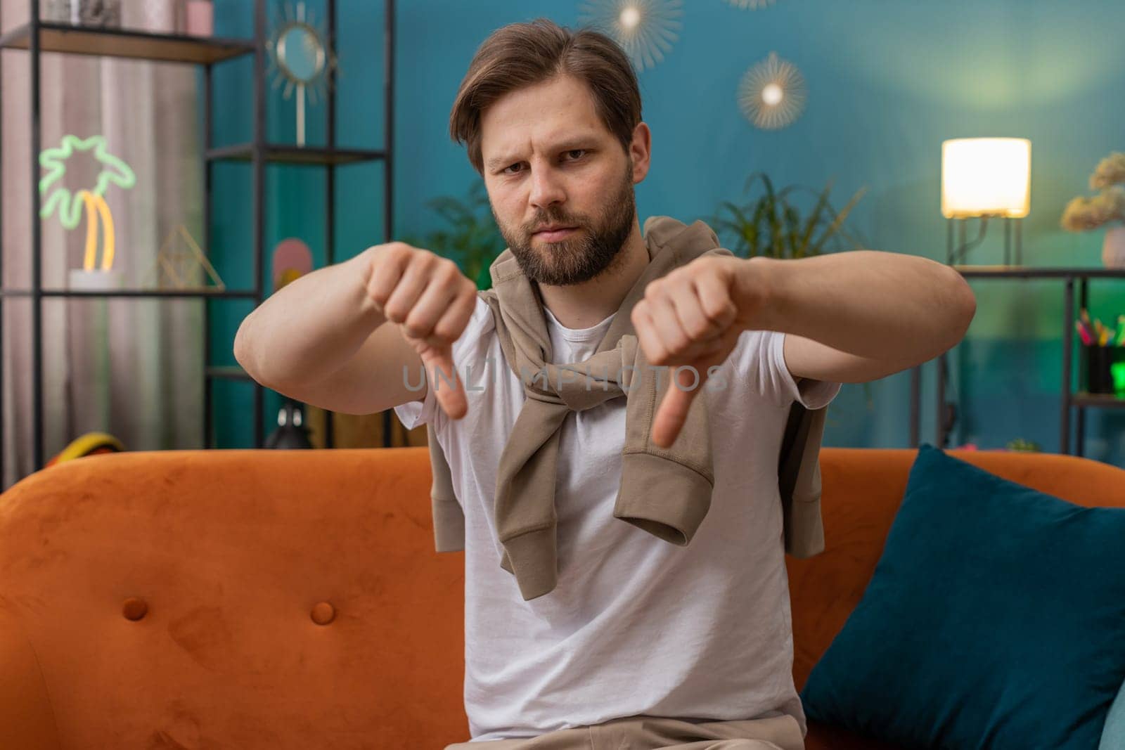 Dislike. Upset young man showing thumbs down sign gesture, expressing discontent, disapproval dissatisfied bad work at modern home apartment indoors. Displeased Caucasian guy in living room on sofa