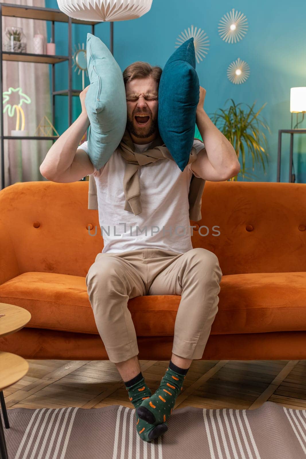 Repair work at neighbours. Irritated young man relaxing on couch cover ears with pillows annoyed by noisy neighbors suffer from headache wish silence. Thin walls at home flat without sound insulation