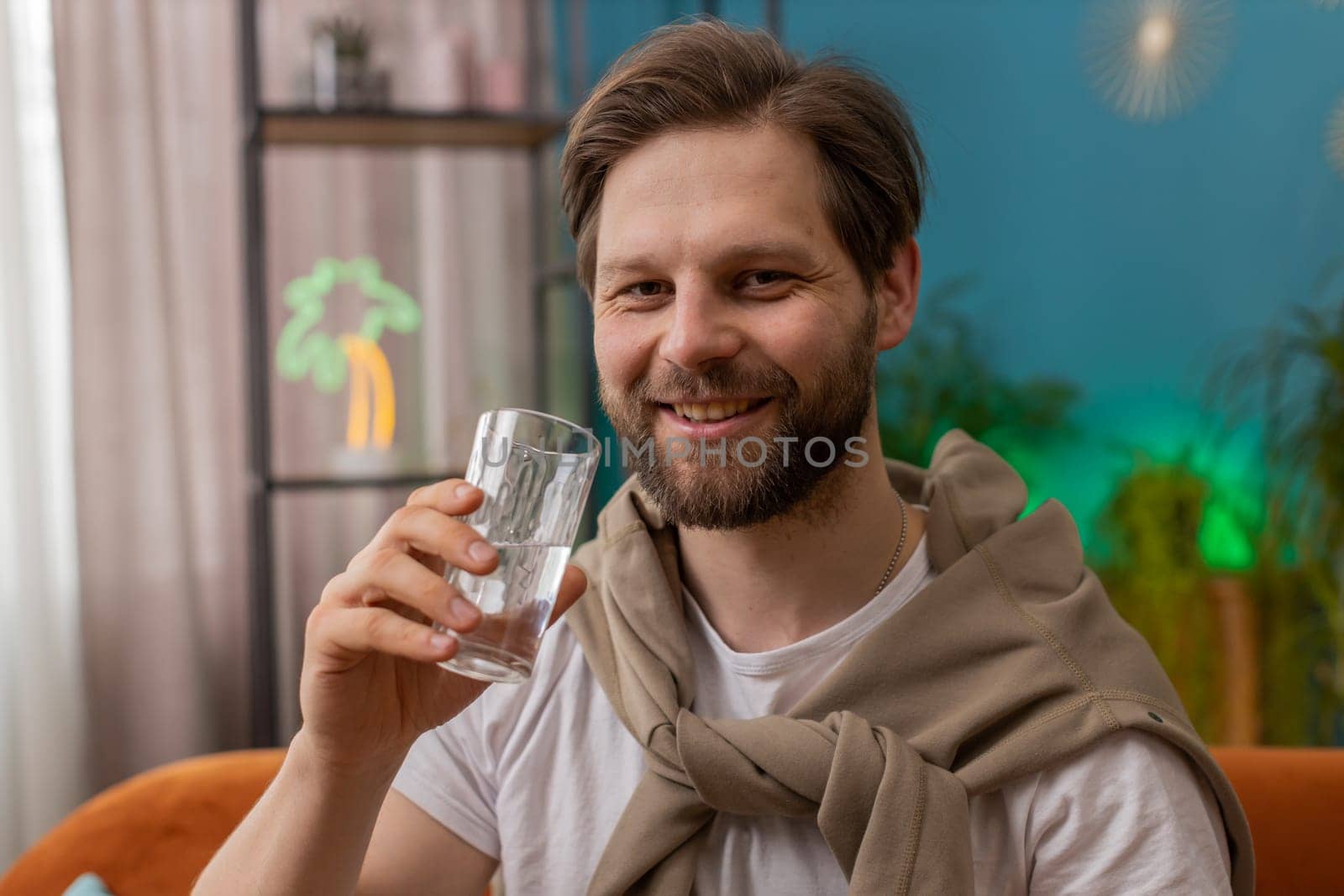 Thirsty man holding glass of natural aqua make sips drinking still water preventing dehydration sits at home living room. Caucasian guy with good life habits, healthy slimming, weight loss concept