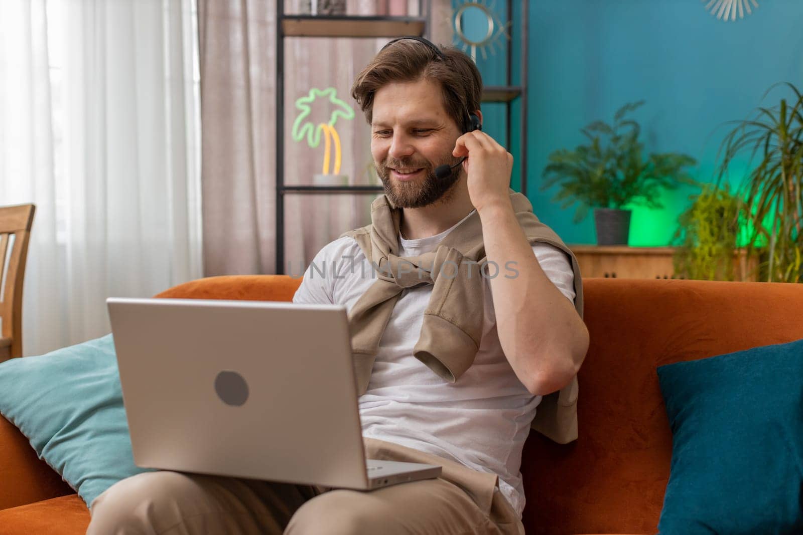 Caucasian man wearing headset, freelance worker, call center or support service operator helpline, having talk with client or colleague communication support waving hello. Guy at home room on sofa