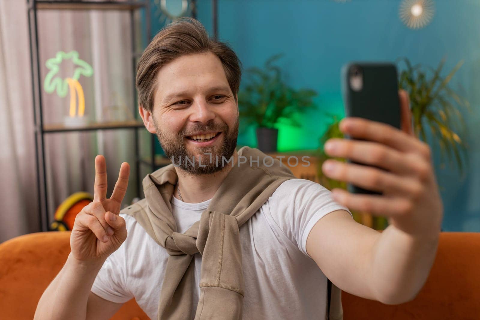 Happy man blogger taking selfie on smartphone, communicating video call online with subscribers, recording stories for social media. Portrait of Caucasian guy at home apartment living room on sofa