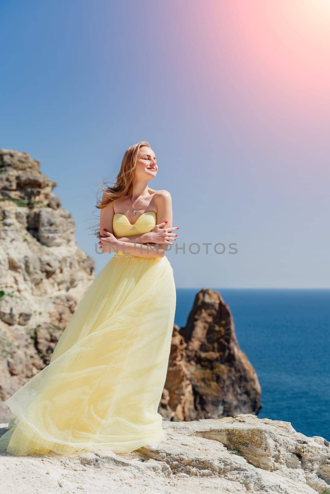 Woman yellow dress sea. Side view Young beautiful sensual woman in yellow long dress posing on a rock high above the sea at sunset. Girl in nature against the blue sky by Matiunina