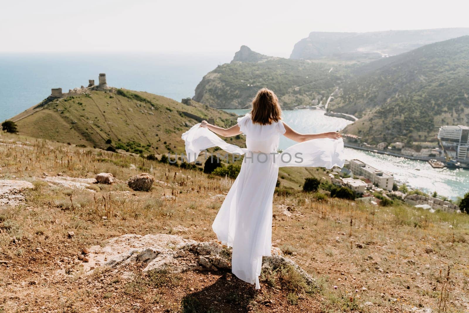 Happy woman in a white dress and hat stands on a rocky cliff above the sea, with the beautiful silhouette of hills in thick fog in the background. by Matiunina