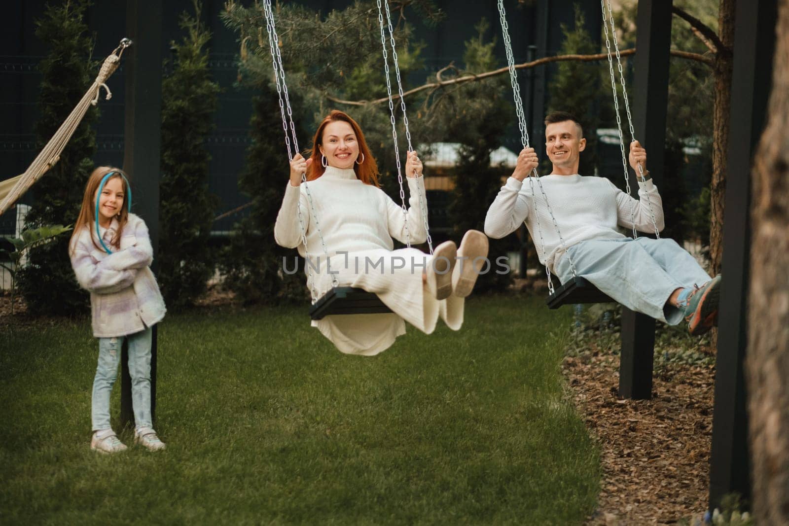 Mom and dad are riding on a swing and there is a daughter standing next to them. The family is resting on a swing.