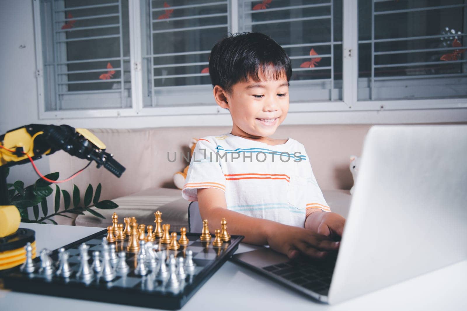 Asian little boy programming code to robot machine arm on laptop for play chess by Sorapop