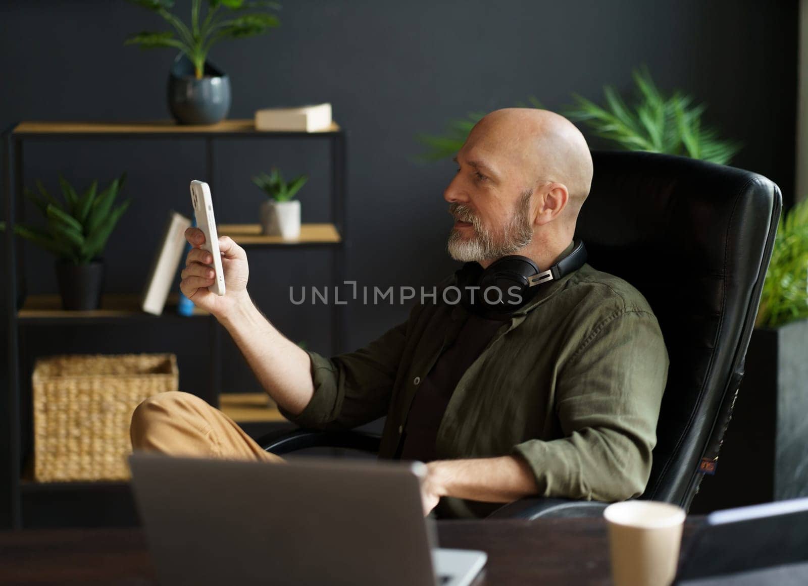 Smart and tech-savvy mid-aged man sits in chair at desk, engrossed in screen of phone. With bald head, silver beard, and air of handsomeness, multitasks, balancing phone, laptop, and cup of coffee. High quality photo
