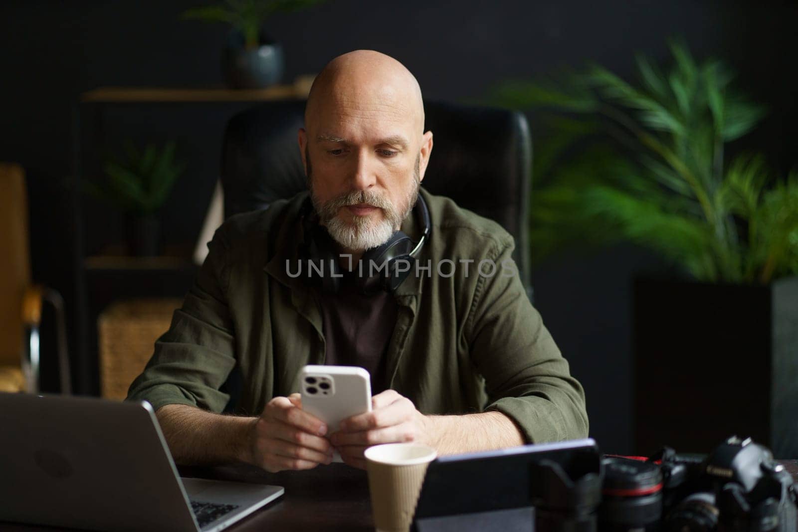 Modern lifestyle of individuals who work remotely or from comfort of own homes. Mature man multitasking at home. He texting message on phone while also working on computer. . High quality photo