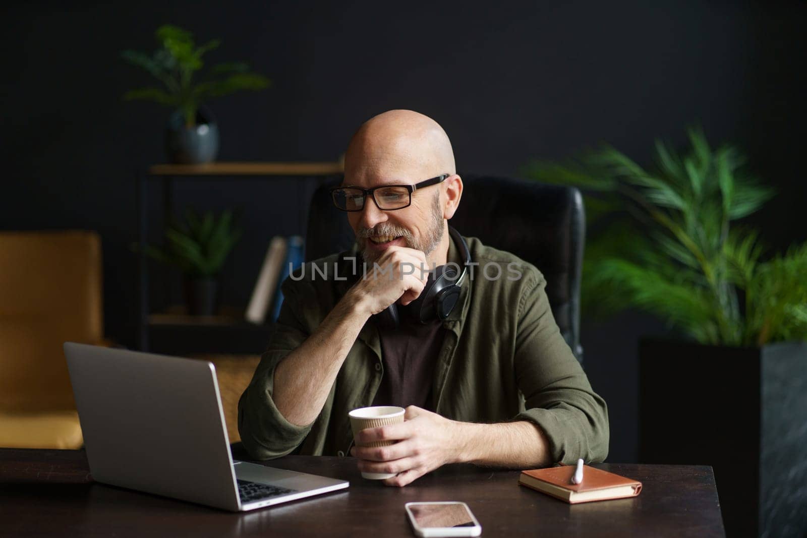 Smiling senior man at home, engrossed in reading news on laptop while holding cup of drink. Enjoying moment of relaxation and staying informed in comfort of his own space. High quality photo
