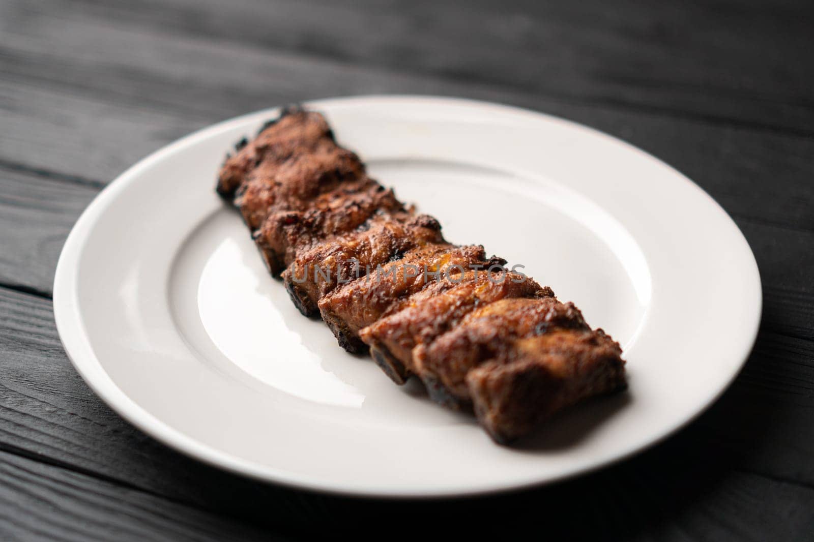 Freshly cooked pork ribs on a white round plate on a black wooden table in the background. Close-up, 45 degree angle view