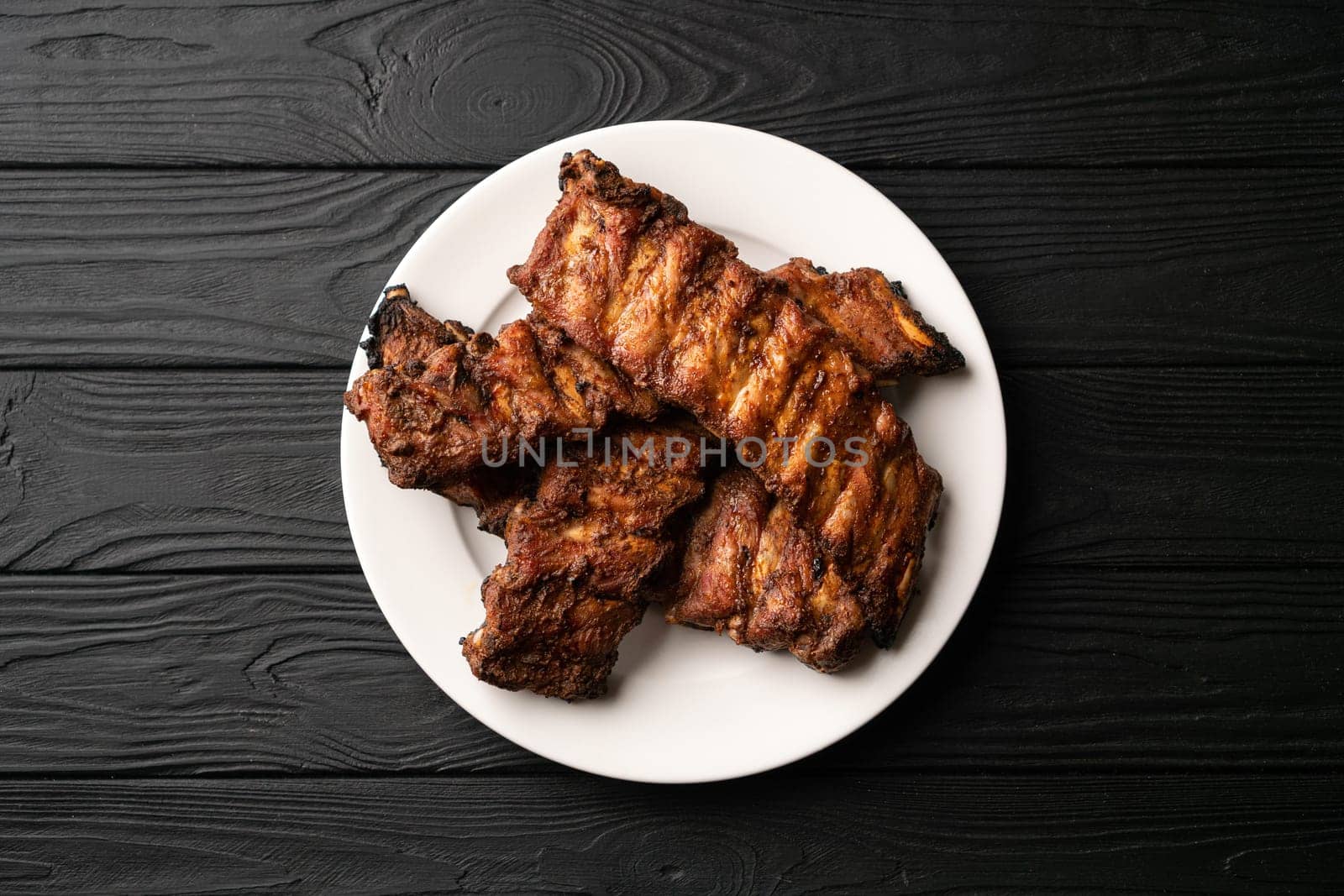 Freshly cooked pork ribs on a white round plate on a black wooden table in the background. Close up, top view